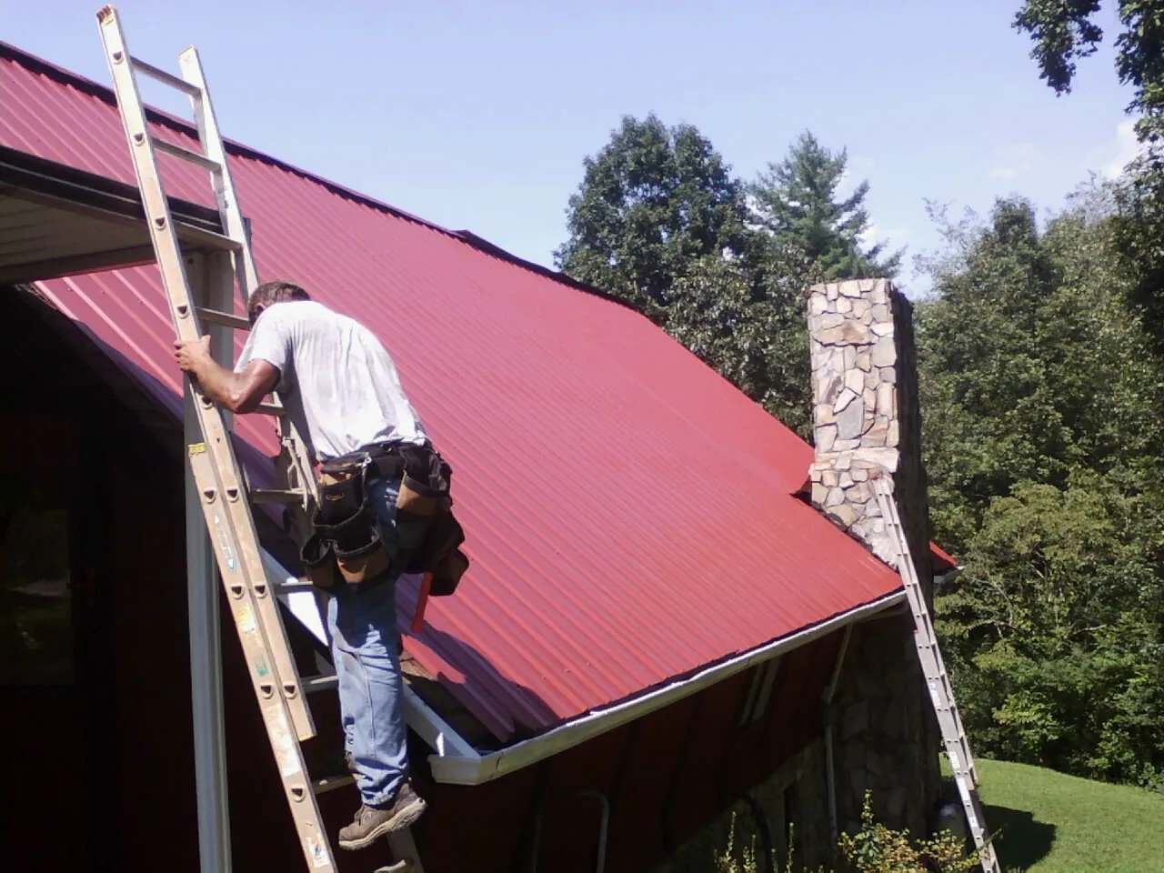 Roofer on a ladder beside a red metal roof and stone chimney — Soffit & Fascia Repair by Roofing Brothers in McCammon